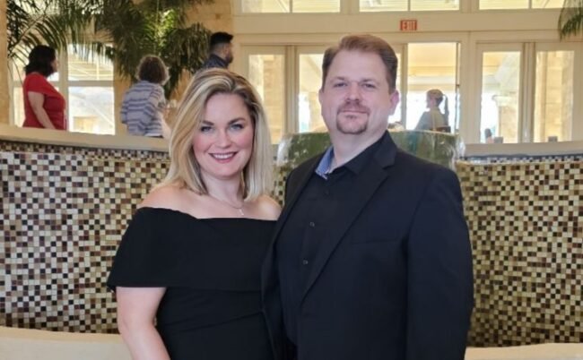A woman and a man posing together indoors, with other people in the background and a tiled wall behind them.