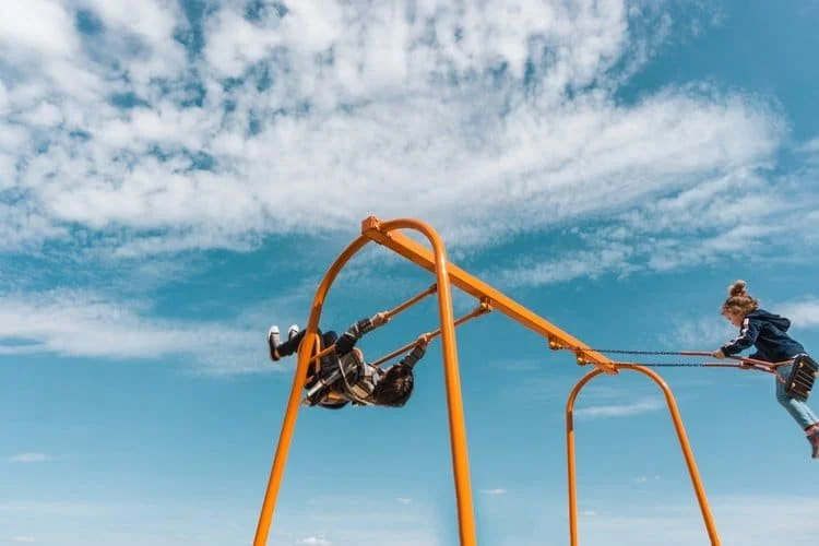 Two children playing on a bright orange swing set outdoors under a blue sky with scattered clouds. One child is swinging upside down, while the other is pushing from the side.