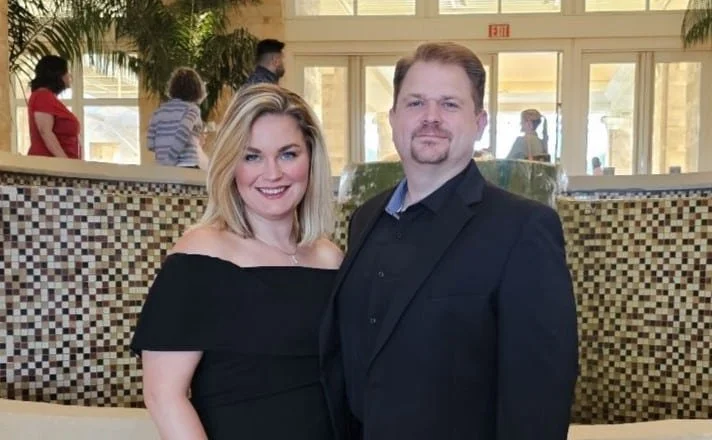 A smiling woman in a black off-the-shoulder dress standing next to a man in a black suit and shirt in a hotel lobby or reception area.