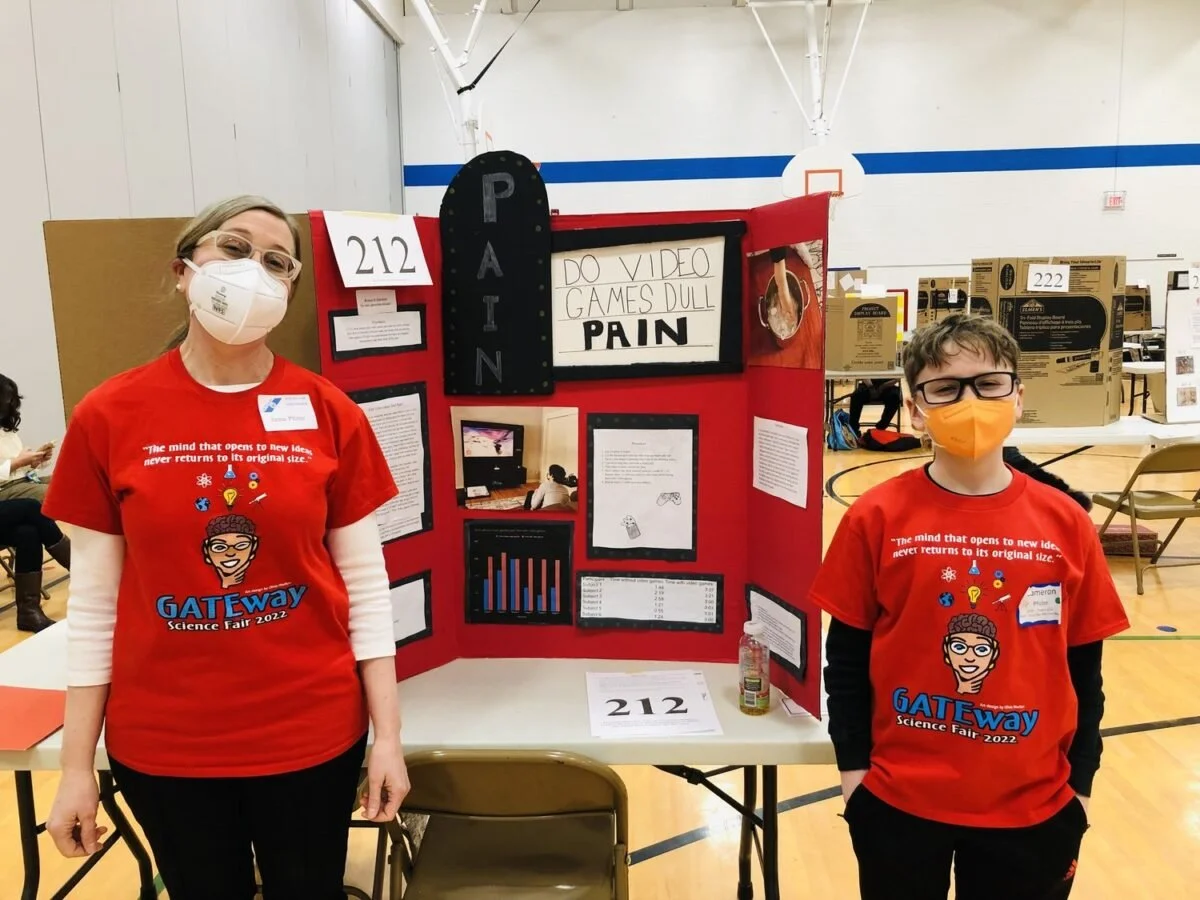 Two children wearing red GATEway Science Fair 2022 shirts and face masks stand in front of a science project display board at a school science fair. The display is titled 'Pain' and includes various posters, graphs, and a framed message about video games.