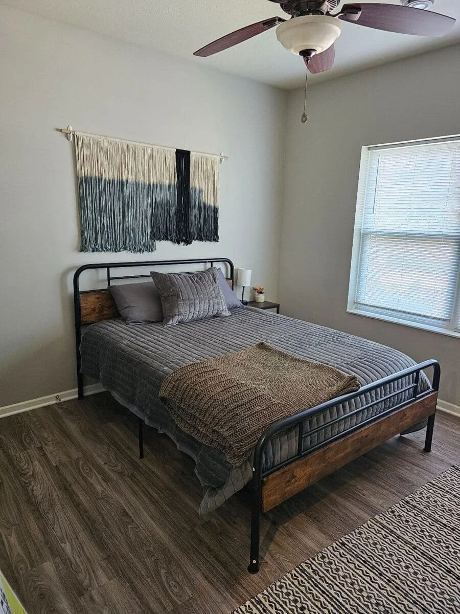 A bedroom with a bed, two pillows, a blanket, a nightstand with a lamp, a window with blinds, a hanging art piece, and a ceiling fan.