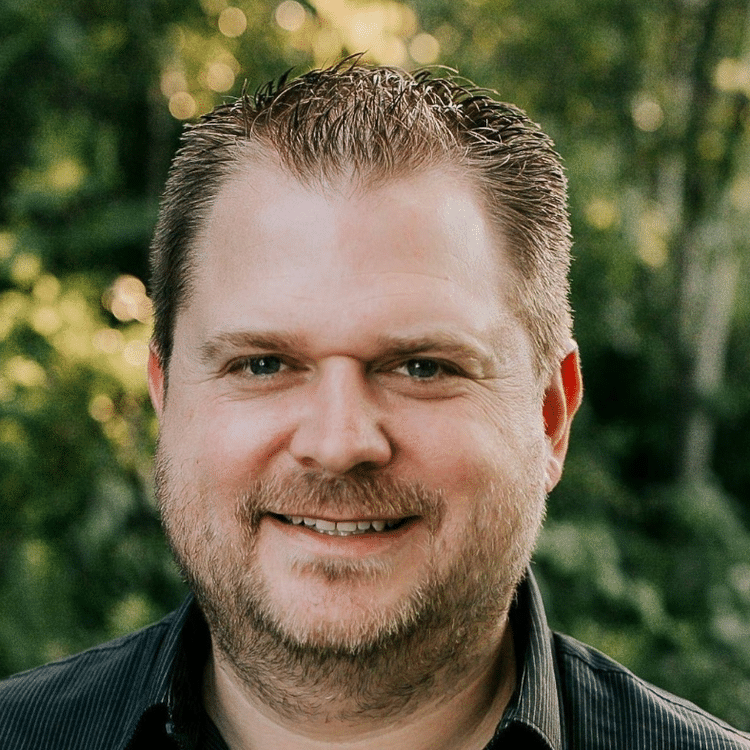 A smiling man with short brown hair and a beard outdoors with green trees in the background.