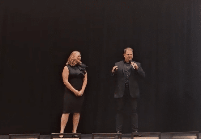 A woman and a man standing on stage, with the man speaking into a microphone and the woman watching him against a black background.