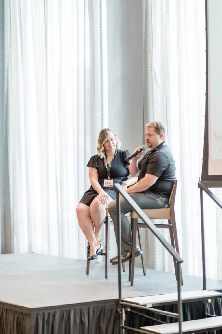 A woman and a man sitting on high chairs on stage at a conference, with the woman holding a microphone to the man, and curtains in the background.