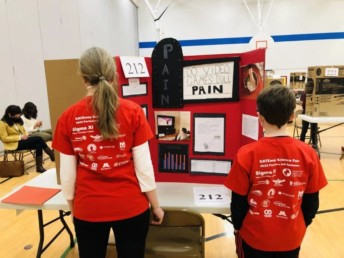 Two students in red T-shirts presenting a science project at a school fair, with a display board titled 'PAIN' and various visual aids, in a gymnasium.