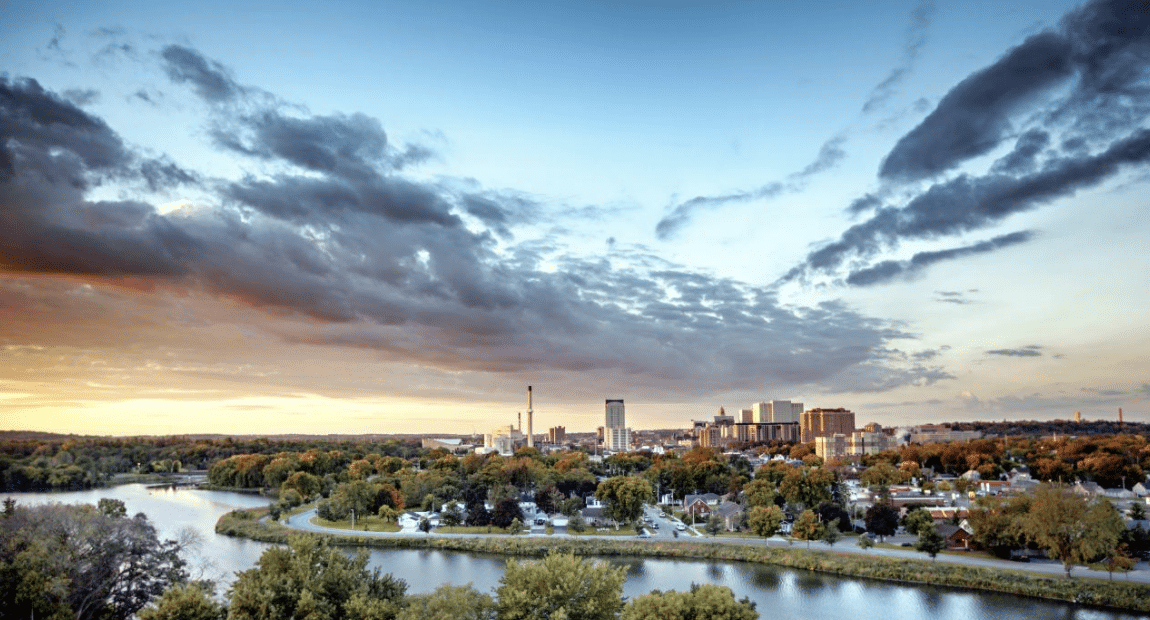 Skyline of a city with a river in the foreground, trees, and clouds overhead during sunset.