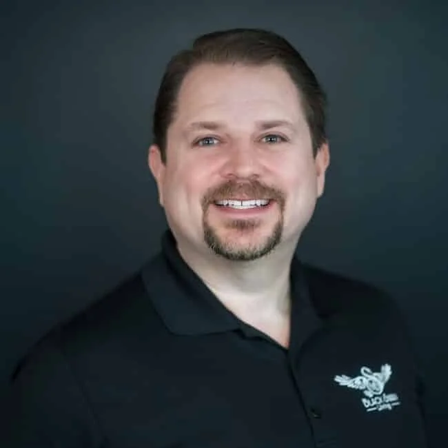 A man with brown hair, a goatee, smiling, wearing a black polo shirt with a logo, against a dark background.