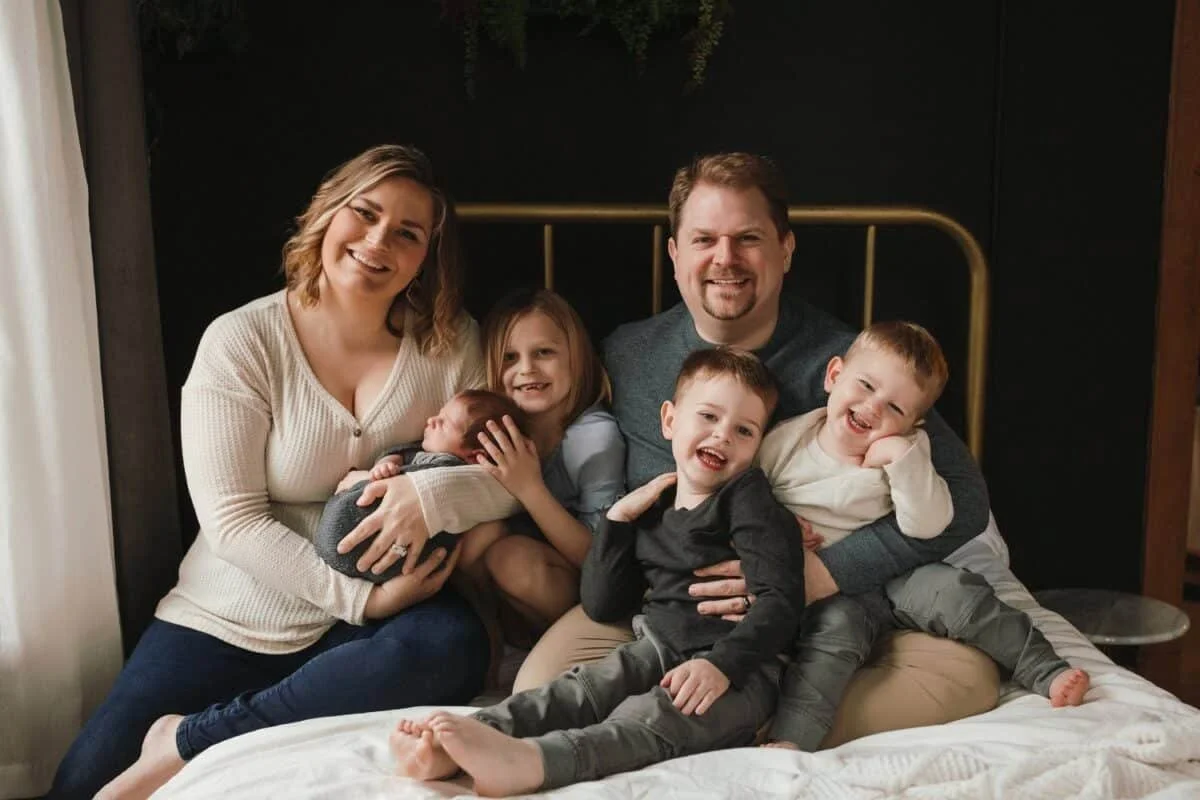 Family of seven sitting on bed, smiling, in a cozy room with black wall and greenery above, including two adults, three young children, and two infants.