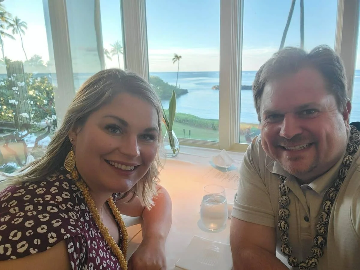 Two people smiling at a restaurant table with a view of the ocean and palm trees outside the window.