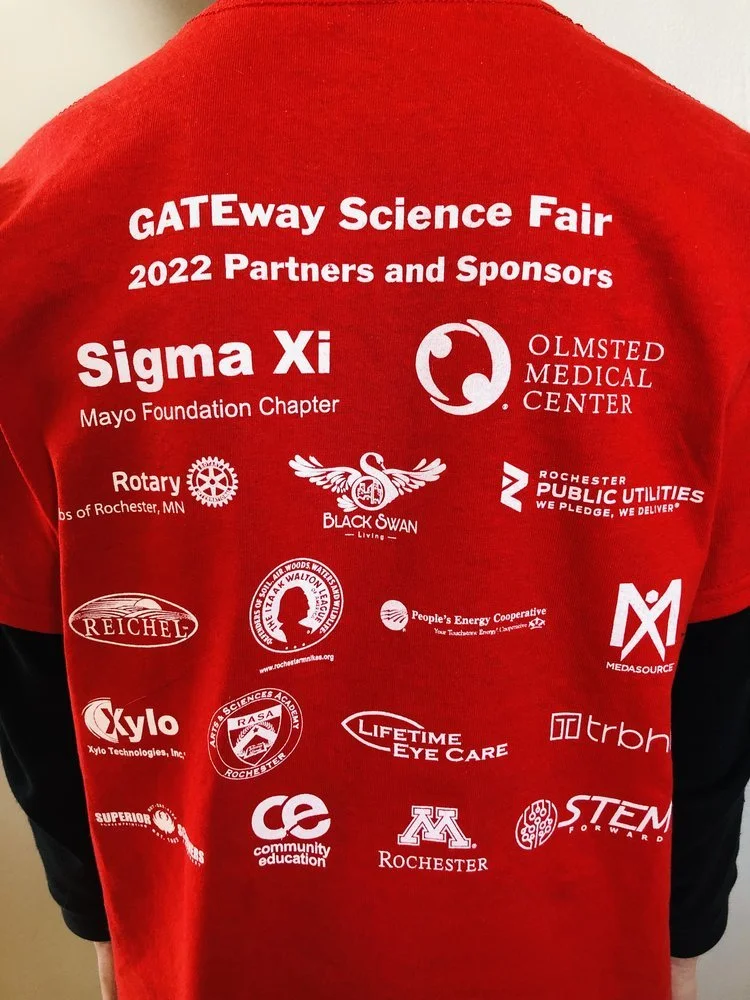 Back of a red shirt with white logos for the GATEway Science Fair 2022 and various sponsors including Sigma Xi, Olmsted Medical Center, Rotary, Black Swan Living, Rochester Public Utilities, Reichel, Rochester Minnesota, Xylo Technologies, Rochester Academy of Science, Lifetime Eye Care, TRBN, Superior, Community Education, University of Minnesota Rochester, and STEM Forward.