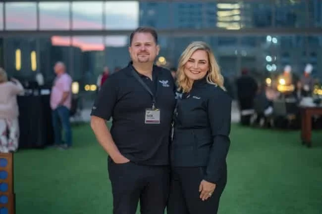 A man and woman standing outdoors at an event, both wearing black shirts with embroidered logos, smiling for the camera.