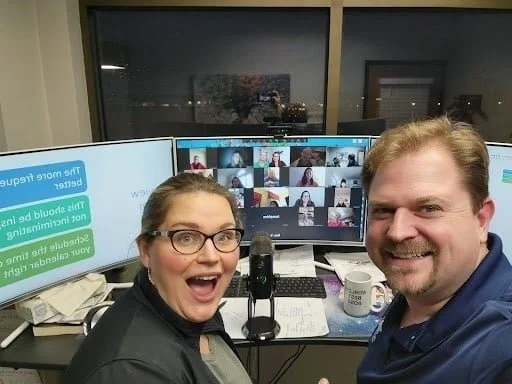 A woman and a man taking a selfie in front of a computer setup with multiple monitors, during a video conference call at night.