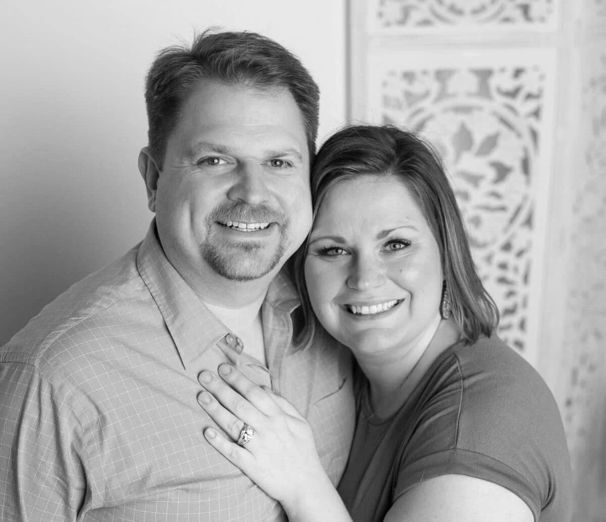 A black-and-white photo of a smiling man and woman embracing each other, with the woman showing an engagement ring.