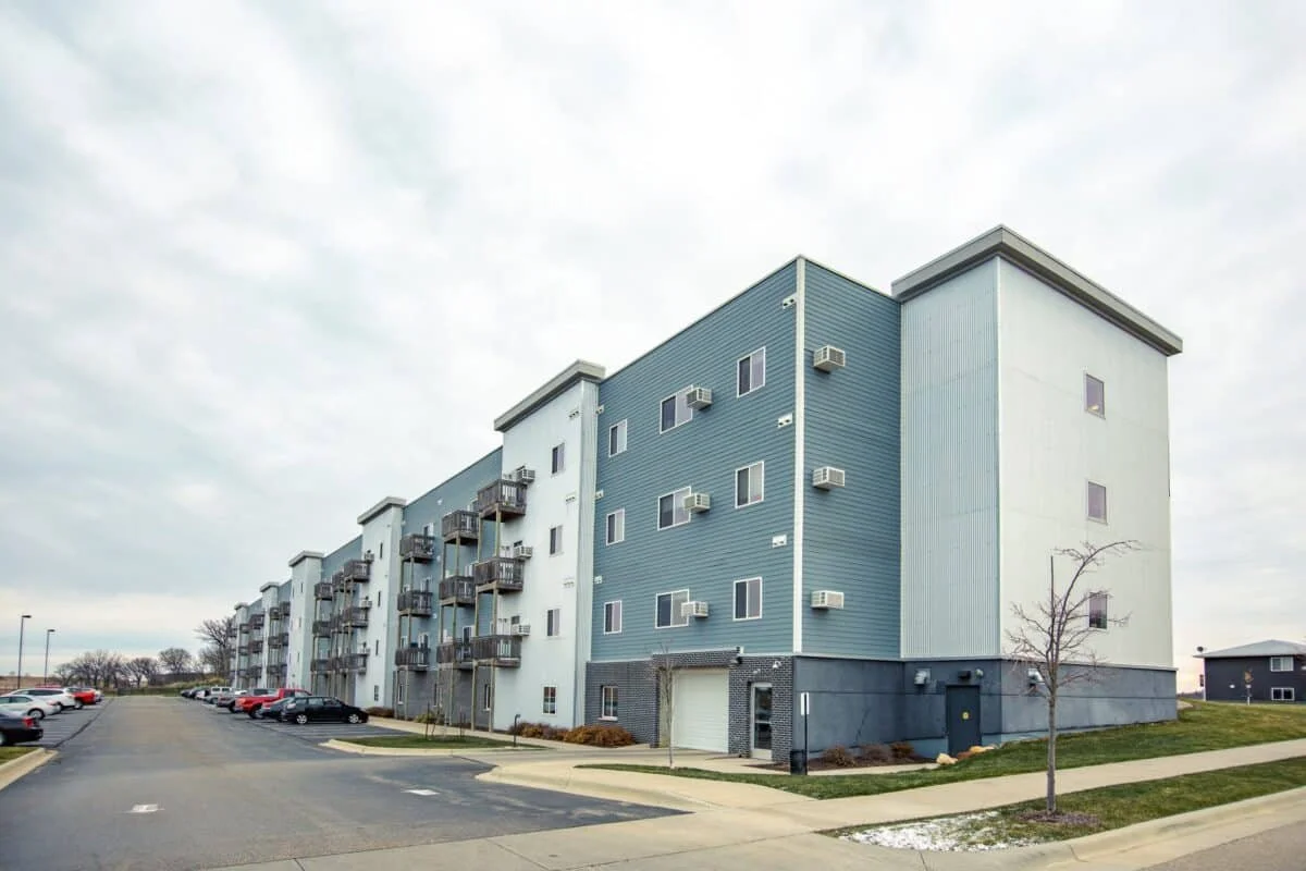 A modern multi-story apartment building with small balconies, parked cars, and a sidewalk with a few trees in a suburban area.
