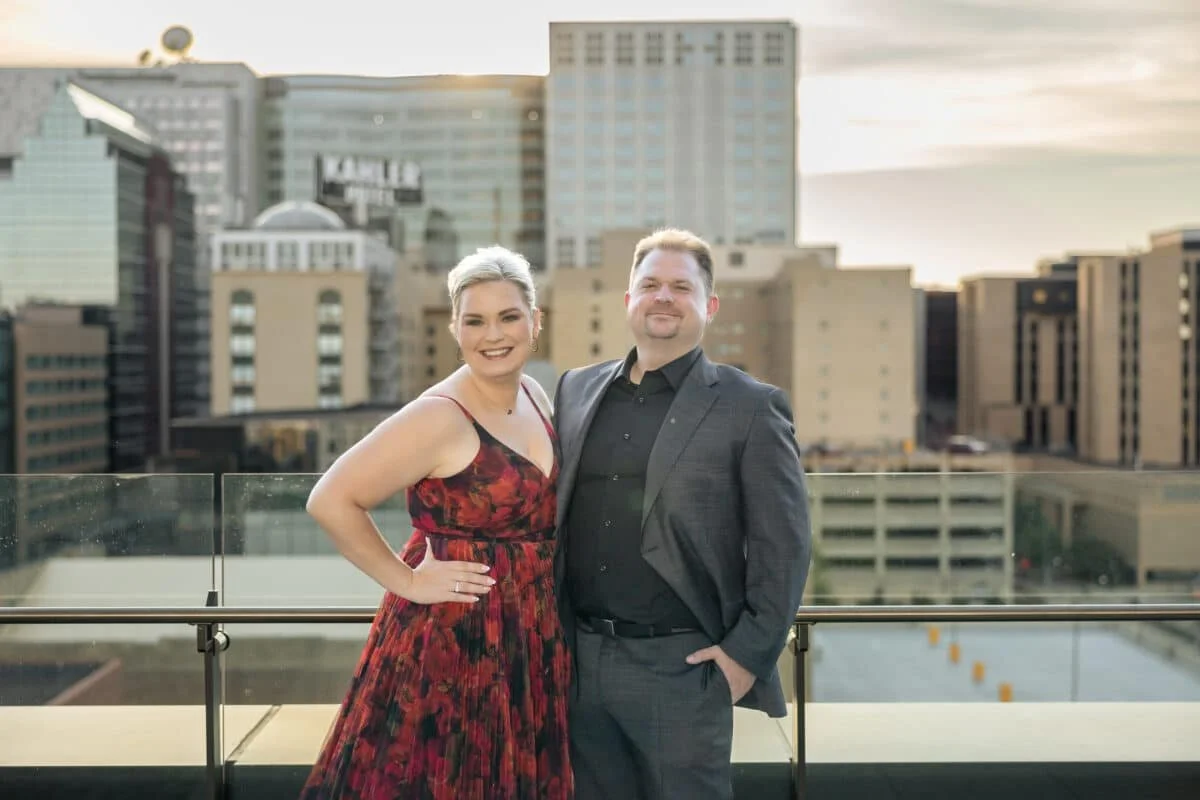 A smiling couple standing on a rooftop with a city skyline in the background during sunset. The woman is wearing a red floral dress, and the man is dressed in a dark suit.