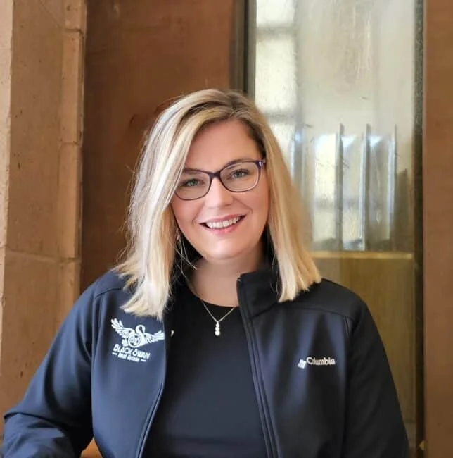 A smiling woman with blonde hair, glasses, and a black jacket standing indoors near a window and brick wall.