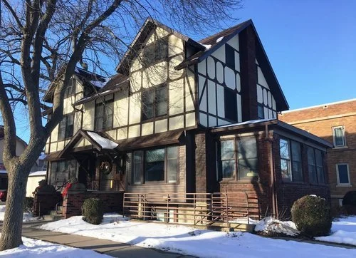 A two-story house with Tudor-style black and white timber framing, partial brick foundation, and a small front porch, set in winter with snow on the ground and leafless trees nearby.