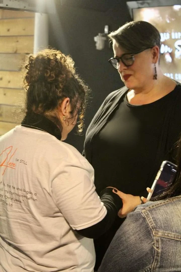 Two women are standing close together, holding hands and praying together. One woman is wearing a white shirt with writing on the back and has curly hair. The other woman, Lisa, is wearing glasses, earrings, and a black shirt, and has short hair.