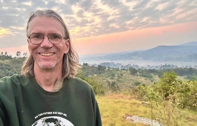 Missionary Tim Way, a man with shoulder-length gray hair, glasses, and a goatee, smiling in front of a scenic landscape in Uganda with hills, trees, and a sky with clouds at sunset or sunrise.