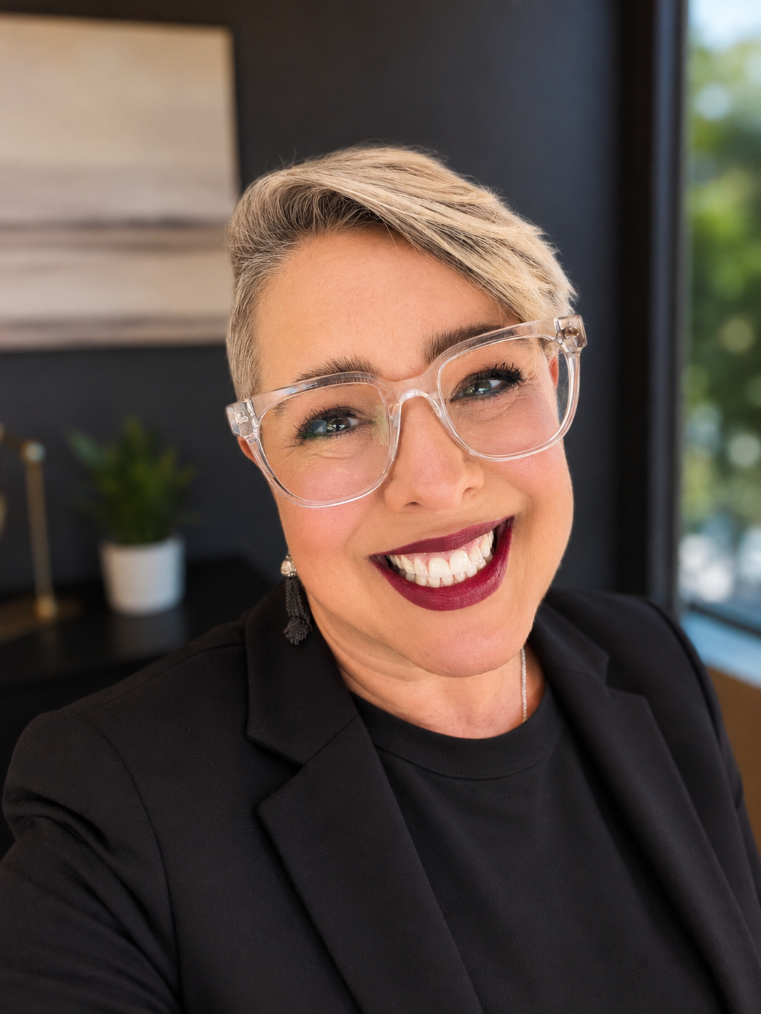 Close-up of Lisa Catalano, a smiling woman with short blonde hair, wearing clear glasses, a black blazer, and black top, in an indoor setting with a blurred background.