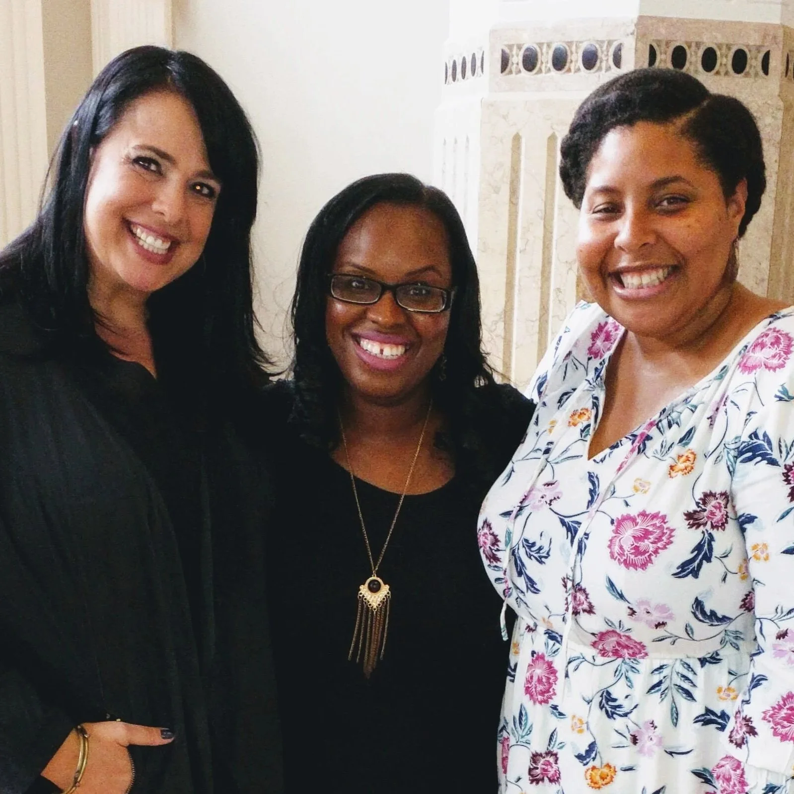 Three women smiling and posing together in a room, standing close to each other. The woman on the left has long dark hair, the woman in the middle has short black hair and glasses, and the woman on the right has textured black hair and is wearing a white dress with a floral pattern.