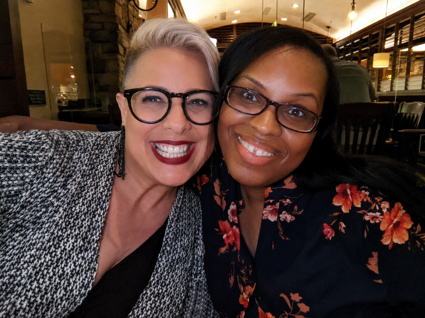 Two women smiling for a close-up photo in a restaurant or cafe, with one woman wearing glasses and a patterned blazer, and the other woman also wearing glasses and a floral shirt.