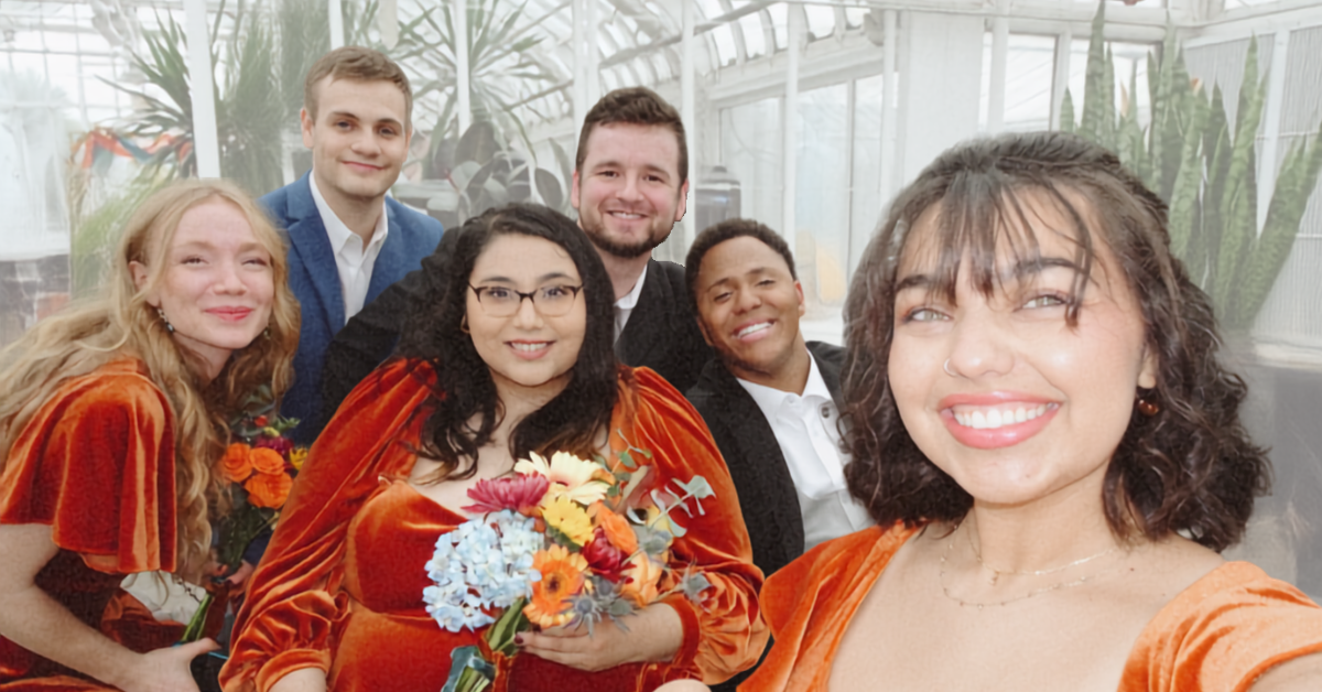 Group of six diverse young adults from Lisa's House Ministries, smiling and posing together at a wedding inside a greenhouse, with some holding flowers, dressed in semi-formal attire with autumn-colored tops.