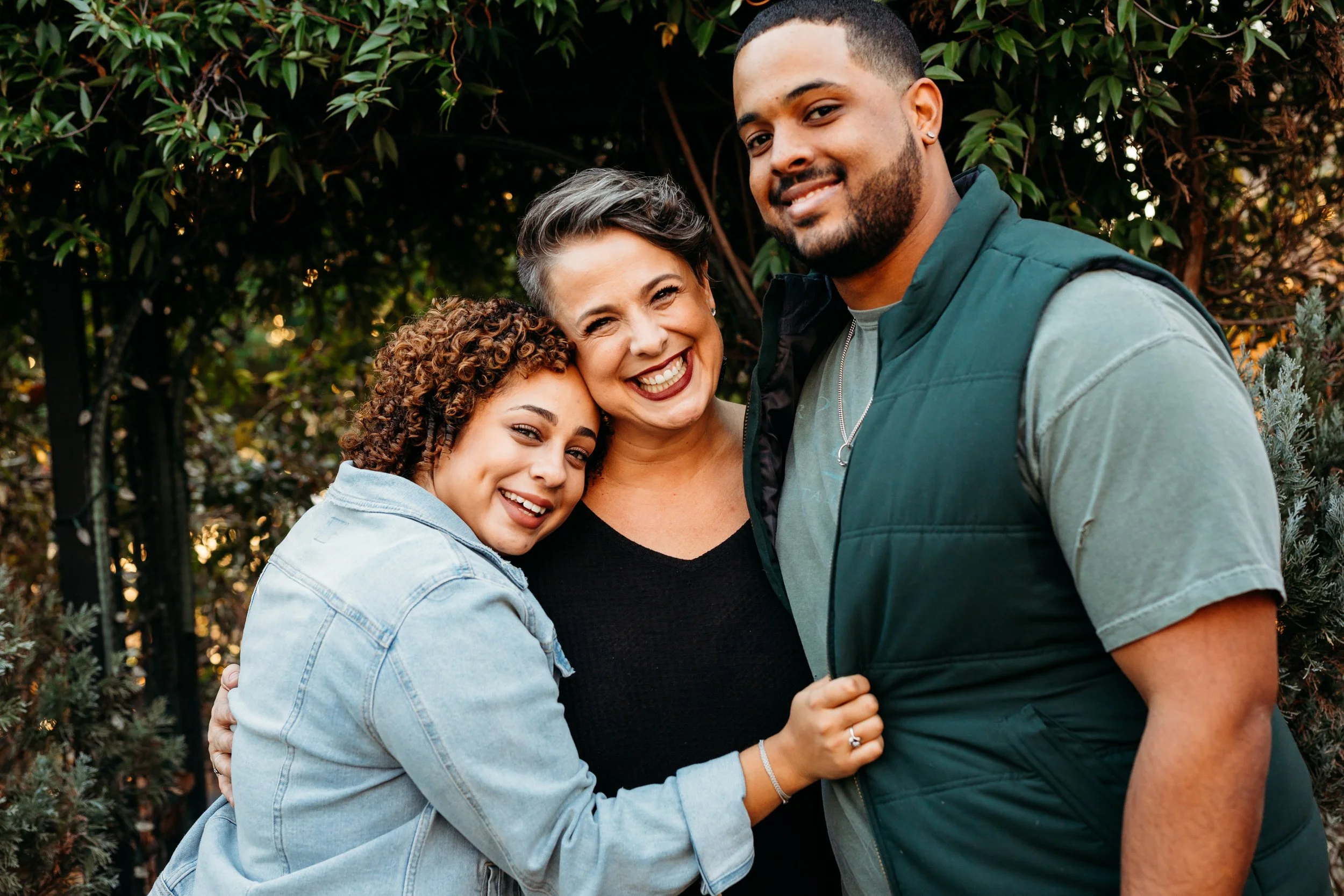 Lisa Catalano and her two adult children. A diverse group of three people smiling and hugging outdoors near greenery.
