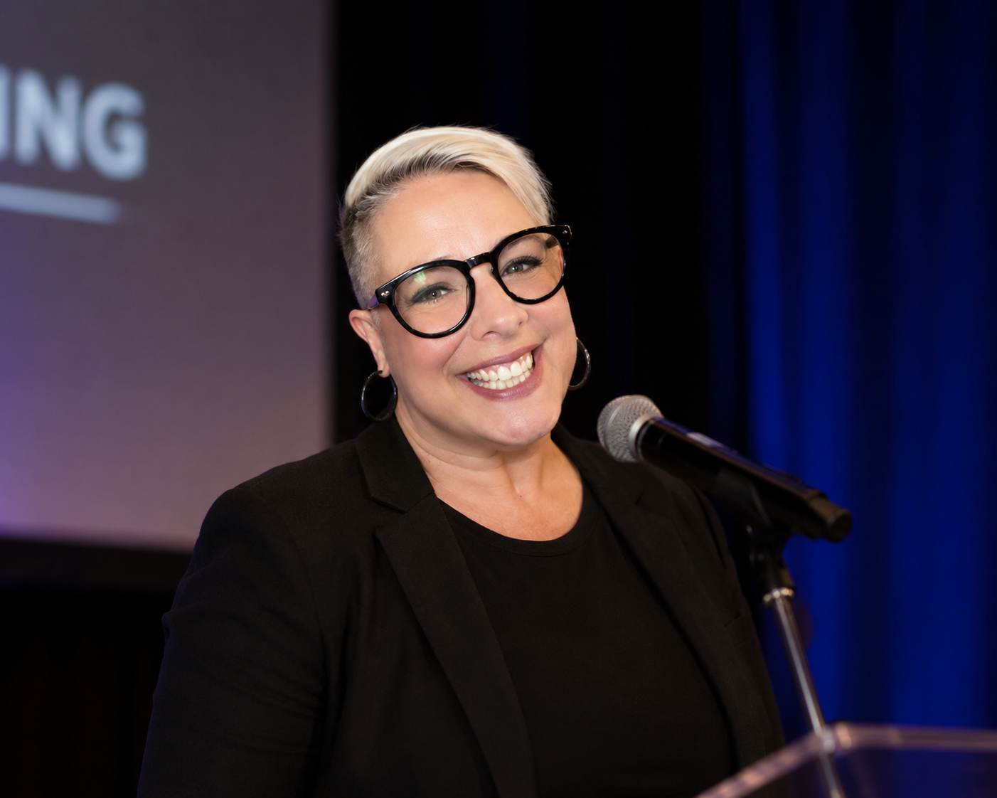 Lisa Catalano, smiling with short blonde hair, glasses, and hoop earrings, speaking at a podium with a microphone.