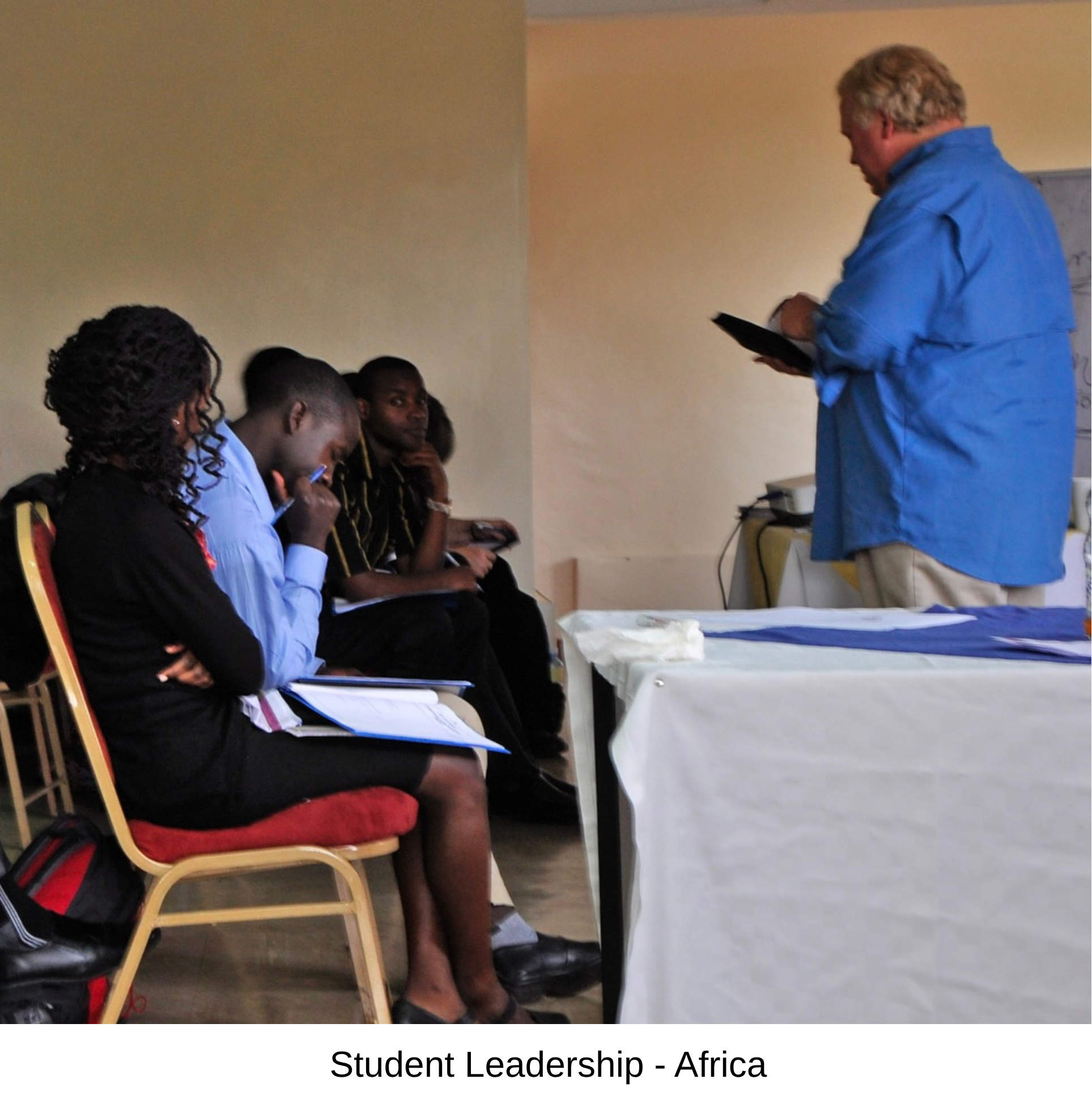 A group of students seated in a classroom, appears to be engaged in a discussion or activity, with a male teacher standing on the right holding a tablet, wearing a blue shirt, labeled as 'Student Leadership - Africa'.