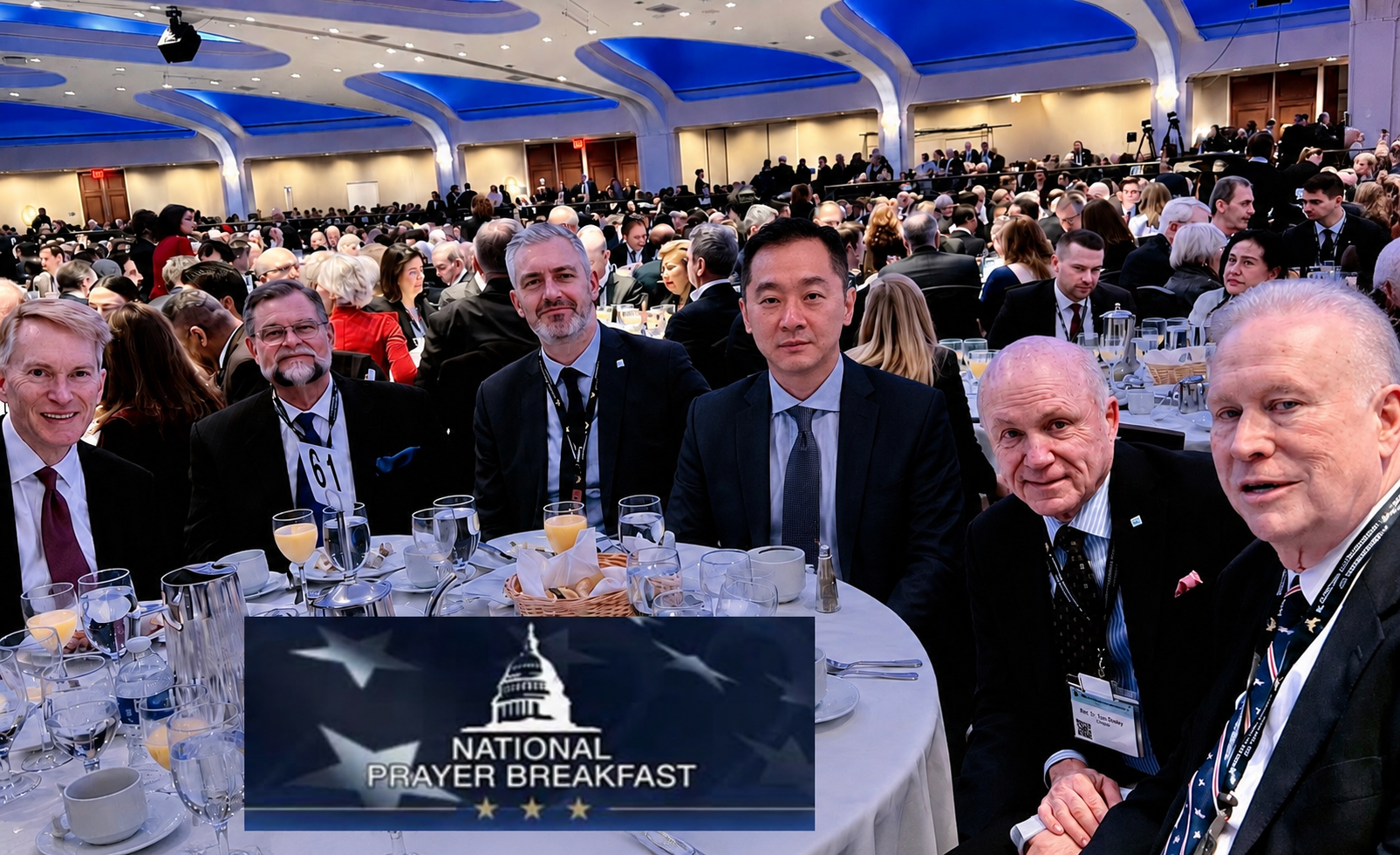Group of six men in suits seated around a table at the National Prayer Breakfast event, with a large crowd in the background and a sign displaying the event logo and name.