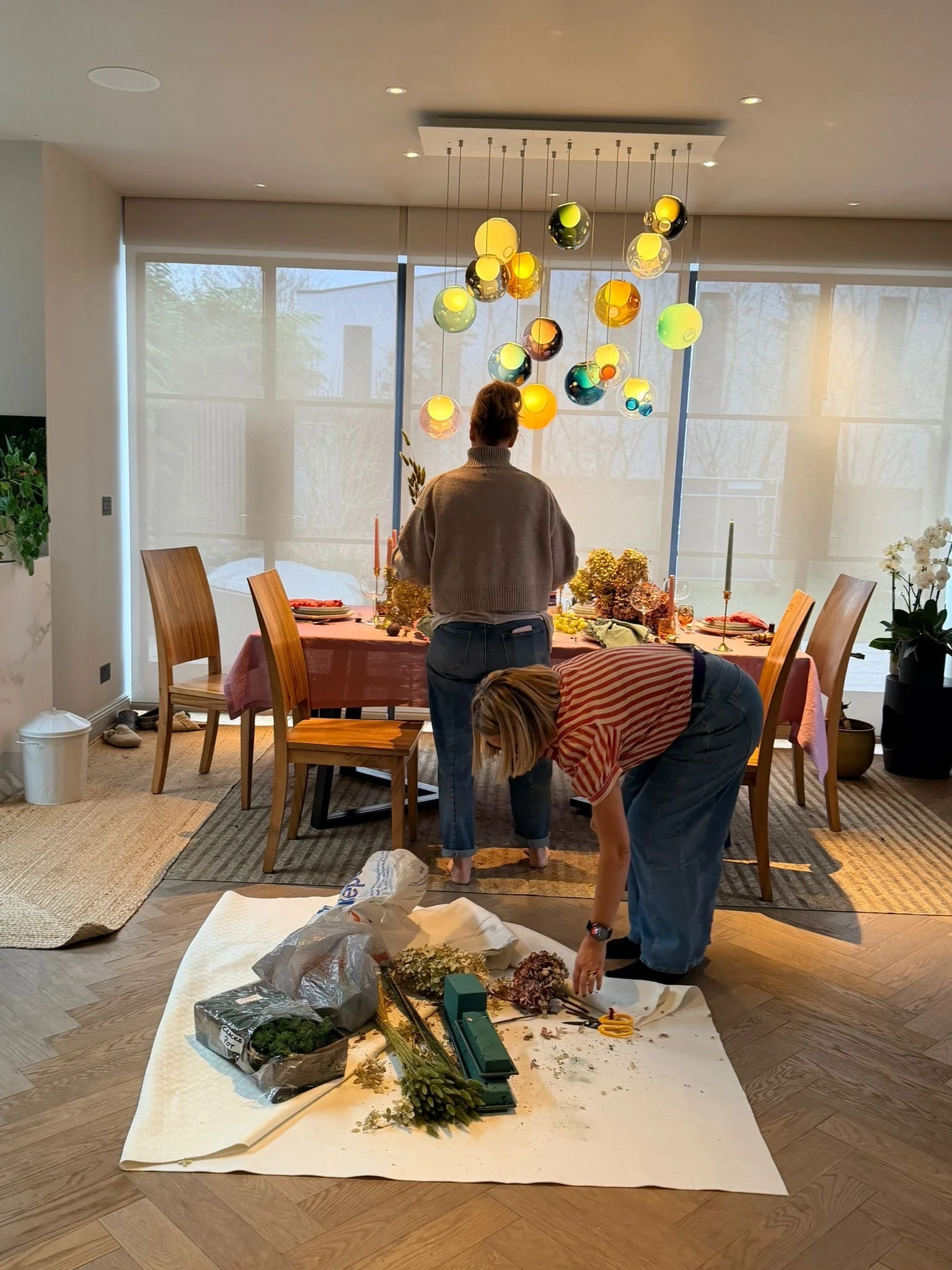 People decorating with flowers and foliage on a dining table, with a large chandelier hanging above, in a bright room with large windows.