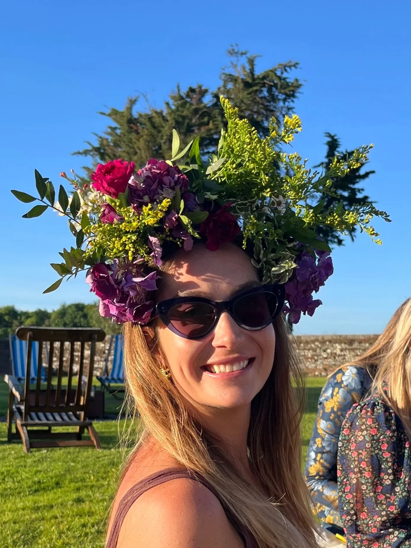 A smiling woman wearing sunglasses and a floral crown made of colorful flowers, outdoors on a sunny day.