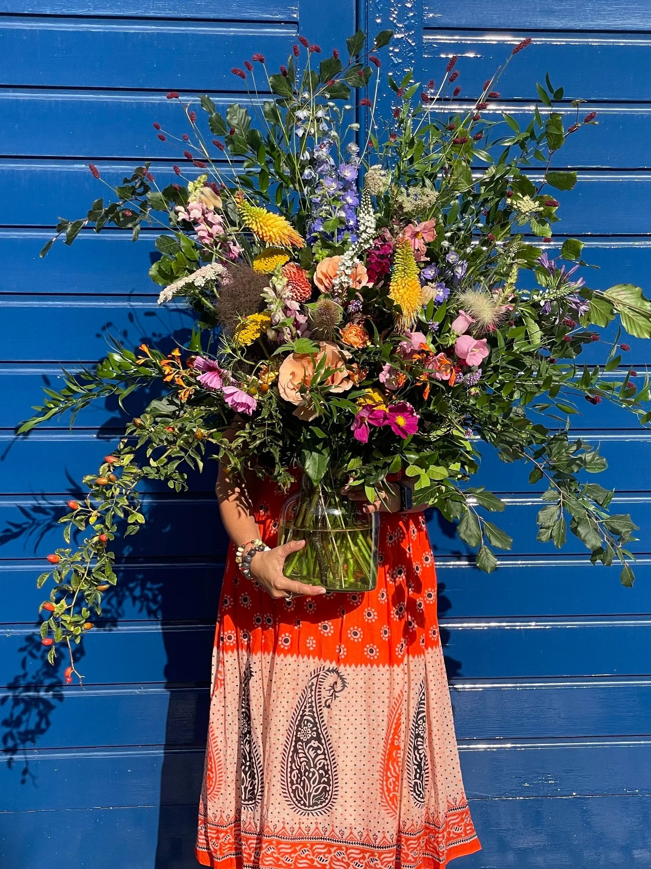 Person holding a large colorful bouquet of flowers in front of a blue wooden wall, wearing a red and orange patterned dress with long sleeves and bracelets.