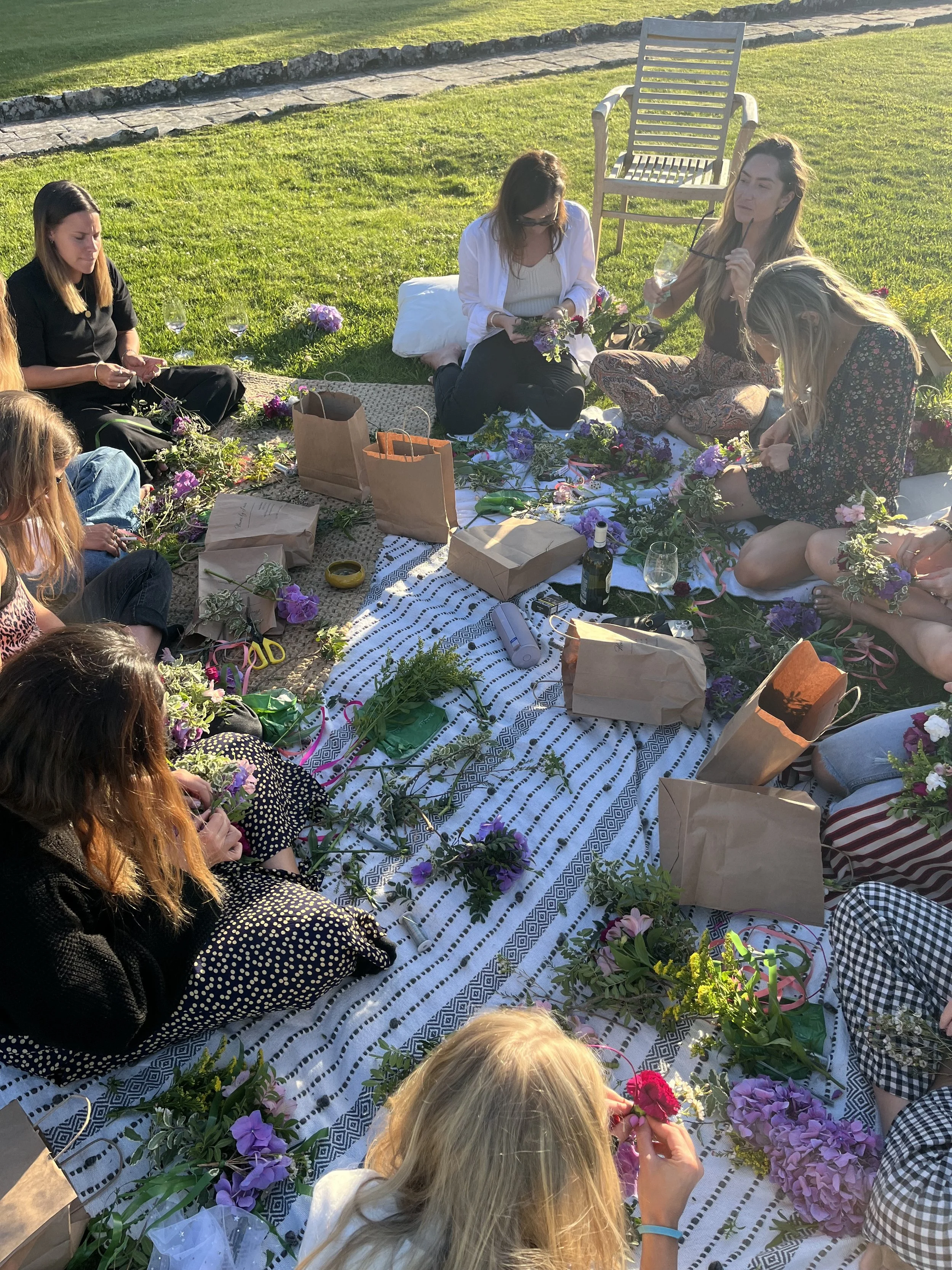 A group of women is sitting and kneeling on a blanket outdoors on a sunny day, surrounded by flowers, with gift bags and wine bottles nearby, possibly preparing floral arrangements.