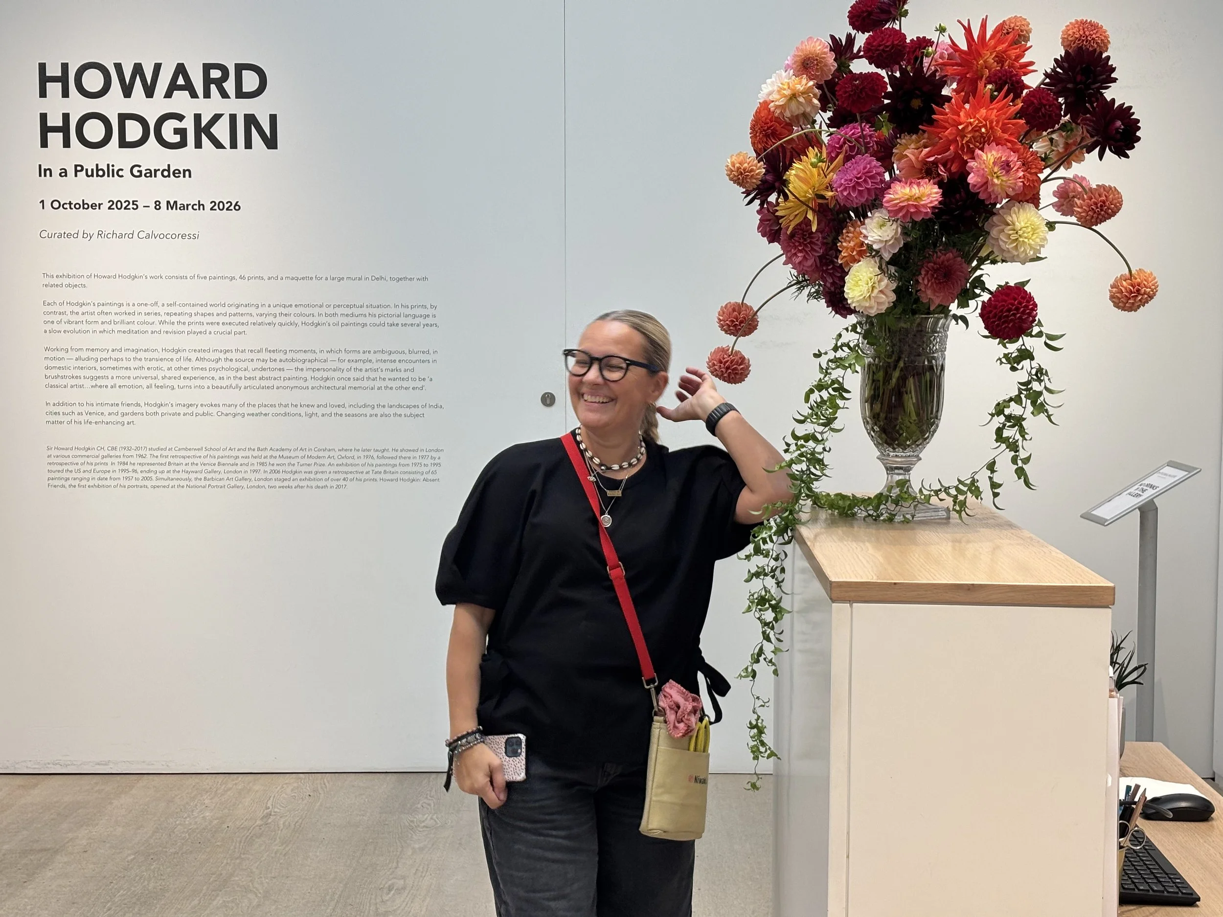 A woman wearing glasses and a black top, smiling and adjusting her hair, standing in front of a large colorful floral arrangement in a gallery or exhibition space.