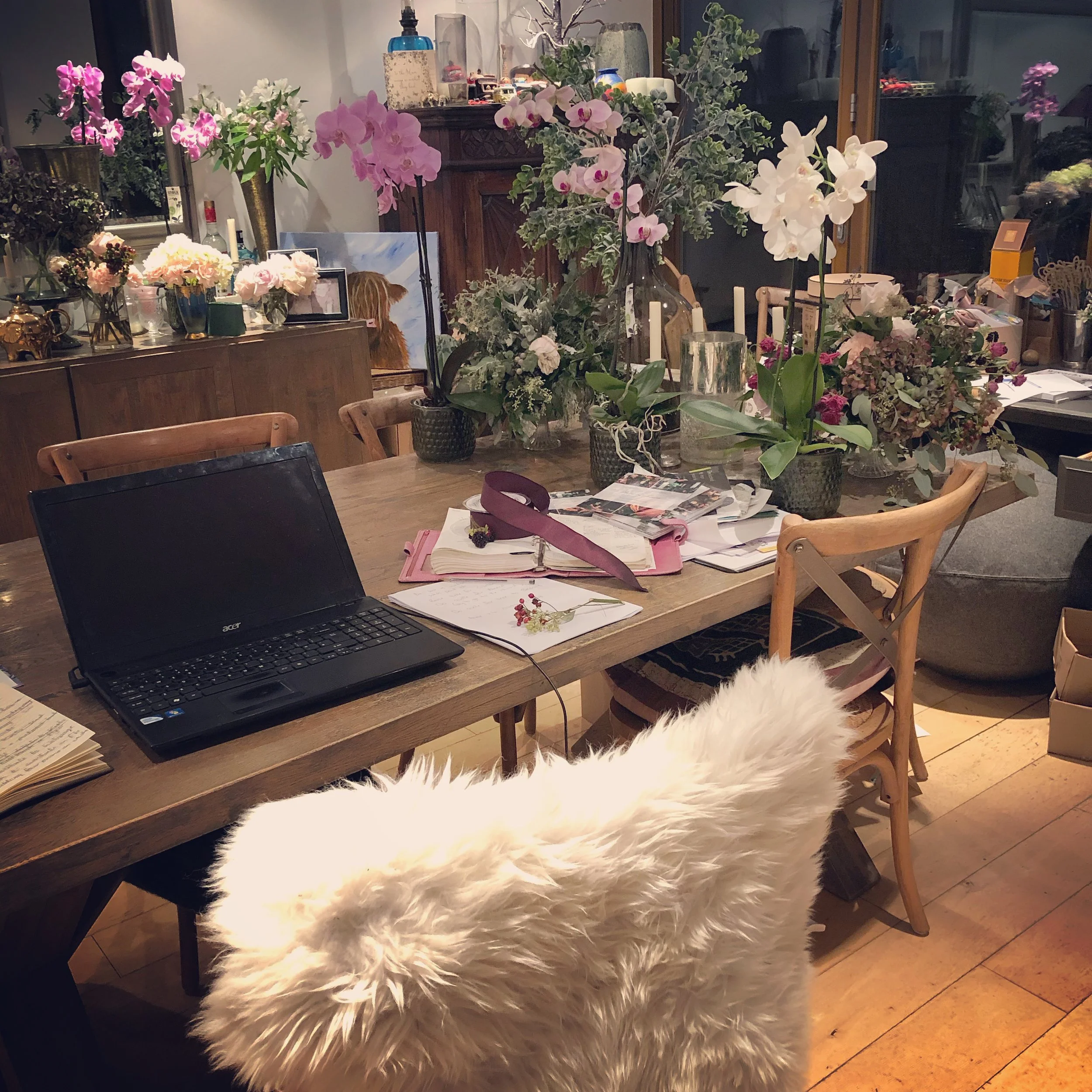 A cozy dining or living room with a table covered in various flowers and plants, a laptop, and papers. In the background, there is a wooden cabinet, vases, and decorative items, with a large window and glass door reflecting the interior."