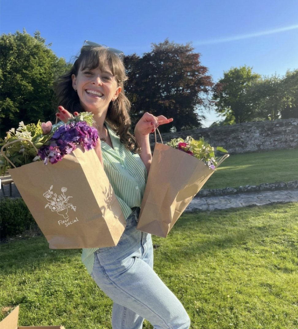 A woman with long brown hair and a big smile holds two paper bags filled with colorful flowers, standing on a grassy outdoor area under a clear blue sky.