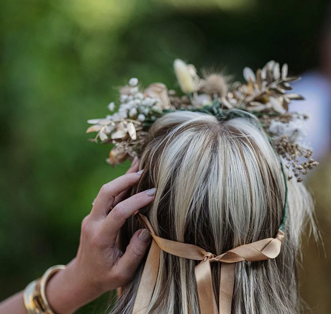 Back of a woman's head with blonde hair, wearing a floral crown and ribbon headband, held by a hand with a gold bracelet, outdoors with a blurred green background.