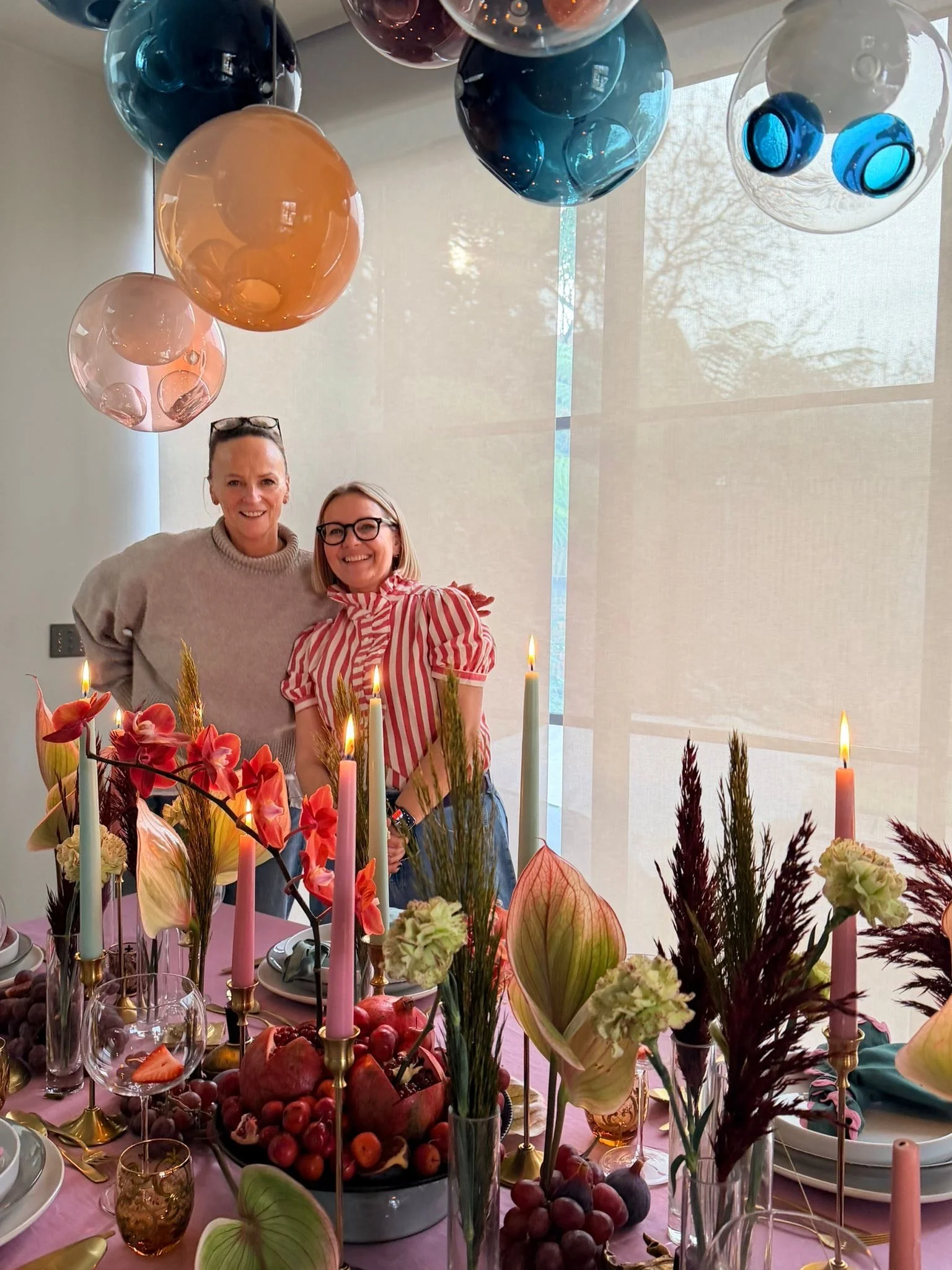 A festive table setting with a variety of flowers, candles, and a large assortment of fruits, including grapes, pomegranates, and apples. In the background, two women are smiling, standing near a window with natural light coming through. Colorful gla