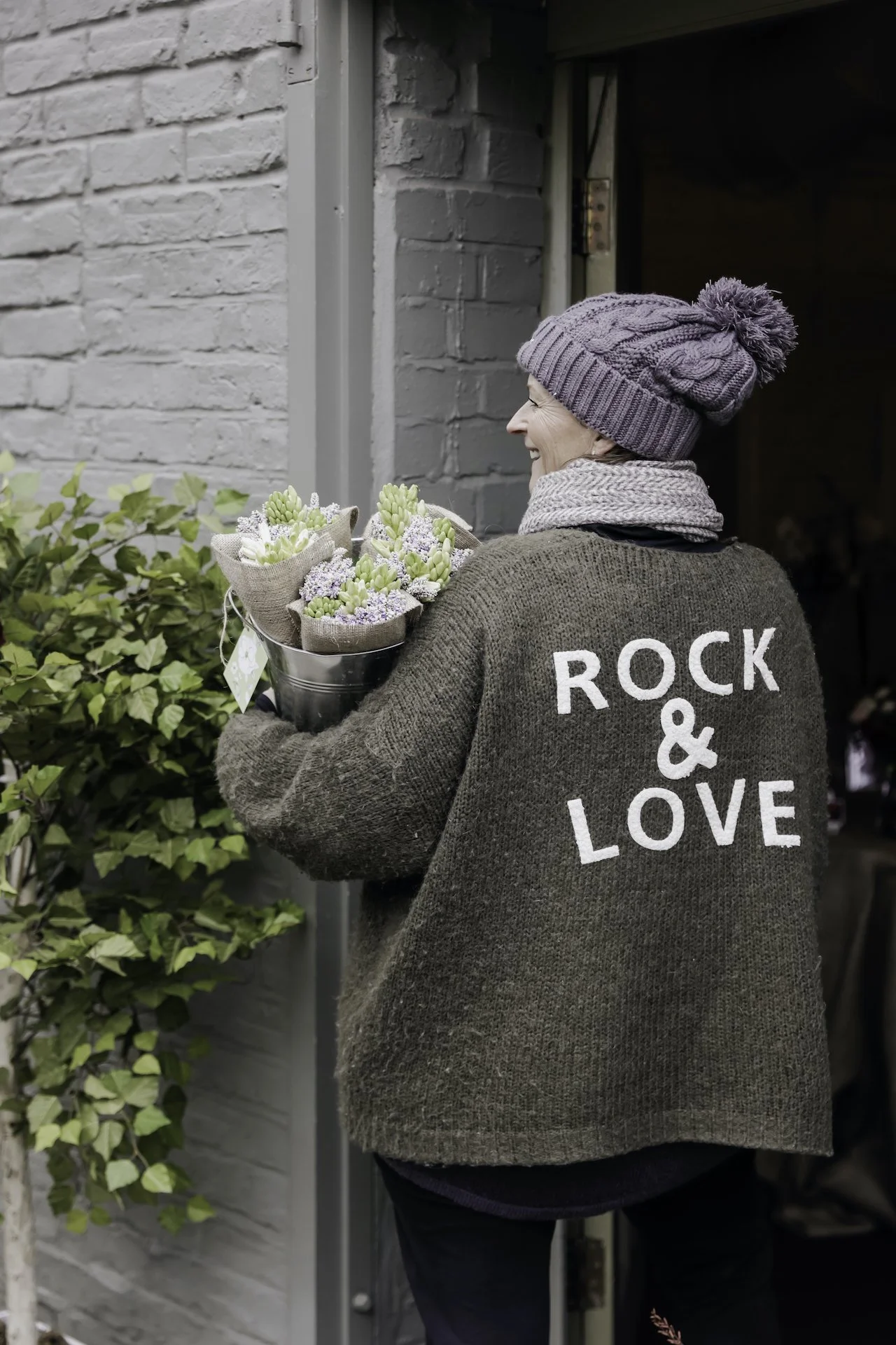 A woman with a gray knit hat and scarf holding a bouquet of flowers in a gray container, standing next to a white brick wall, smiling.