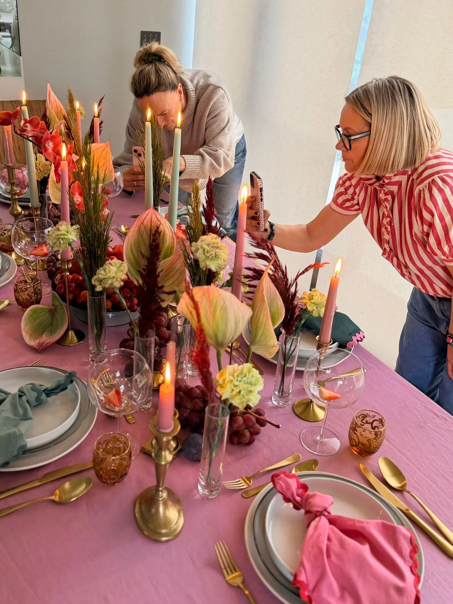 A decorated dining table with pink tablecloth, gold utensils, floral centerpiece, pink candles, wine glasses, and pink and gold tableware, set for a celebration.
