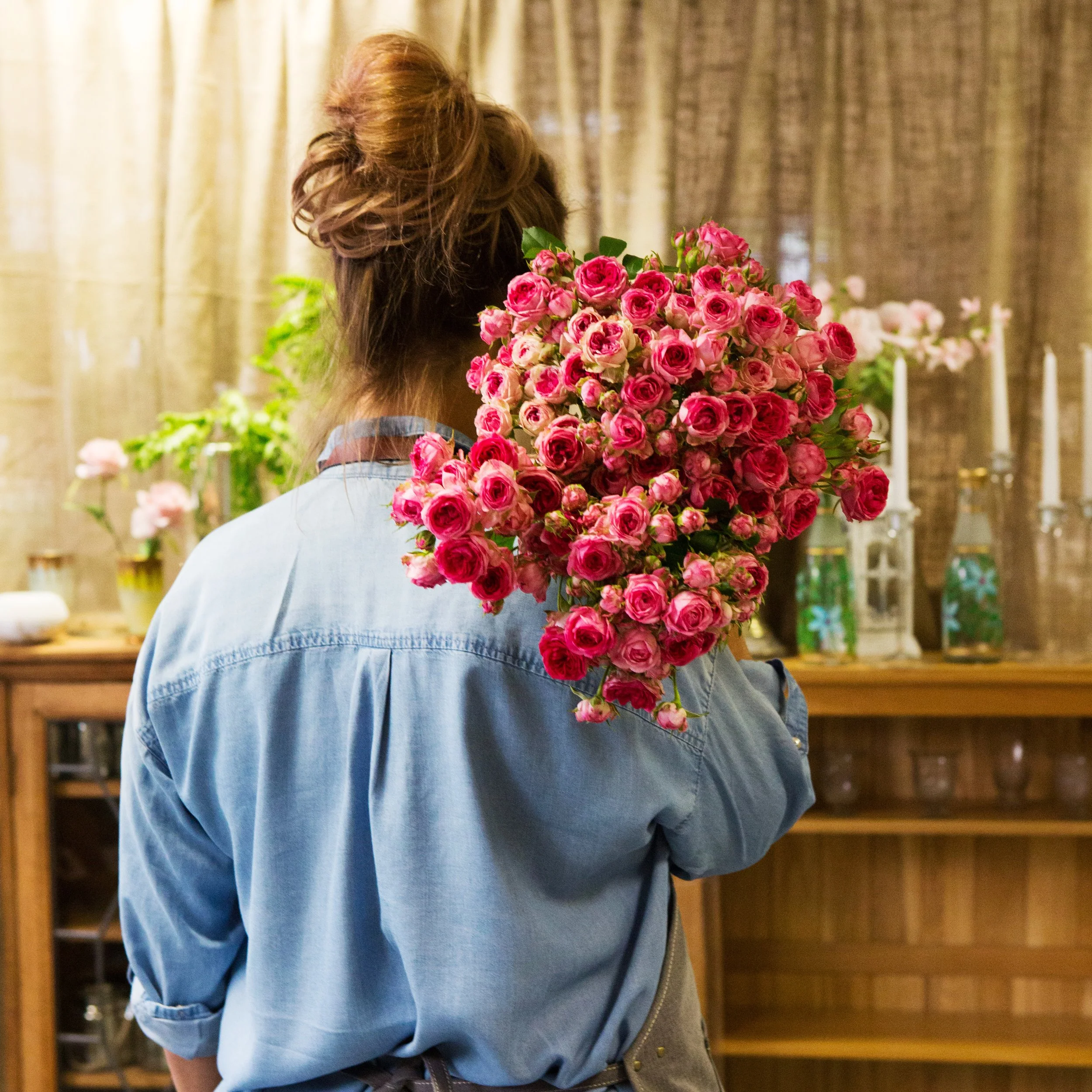 Person with long hair in a bun, wearing a denim jacket, holding a large bouquet of pink roses, standing indoors with a wooden shelf and curtains in the background.