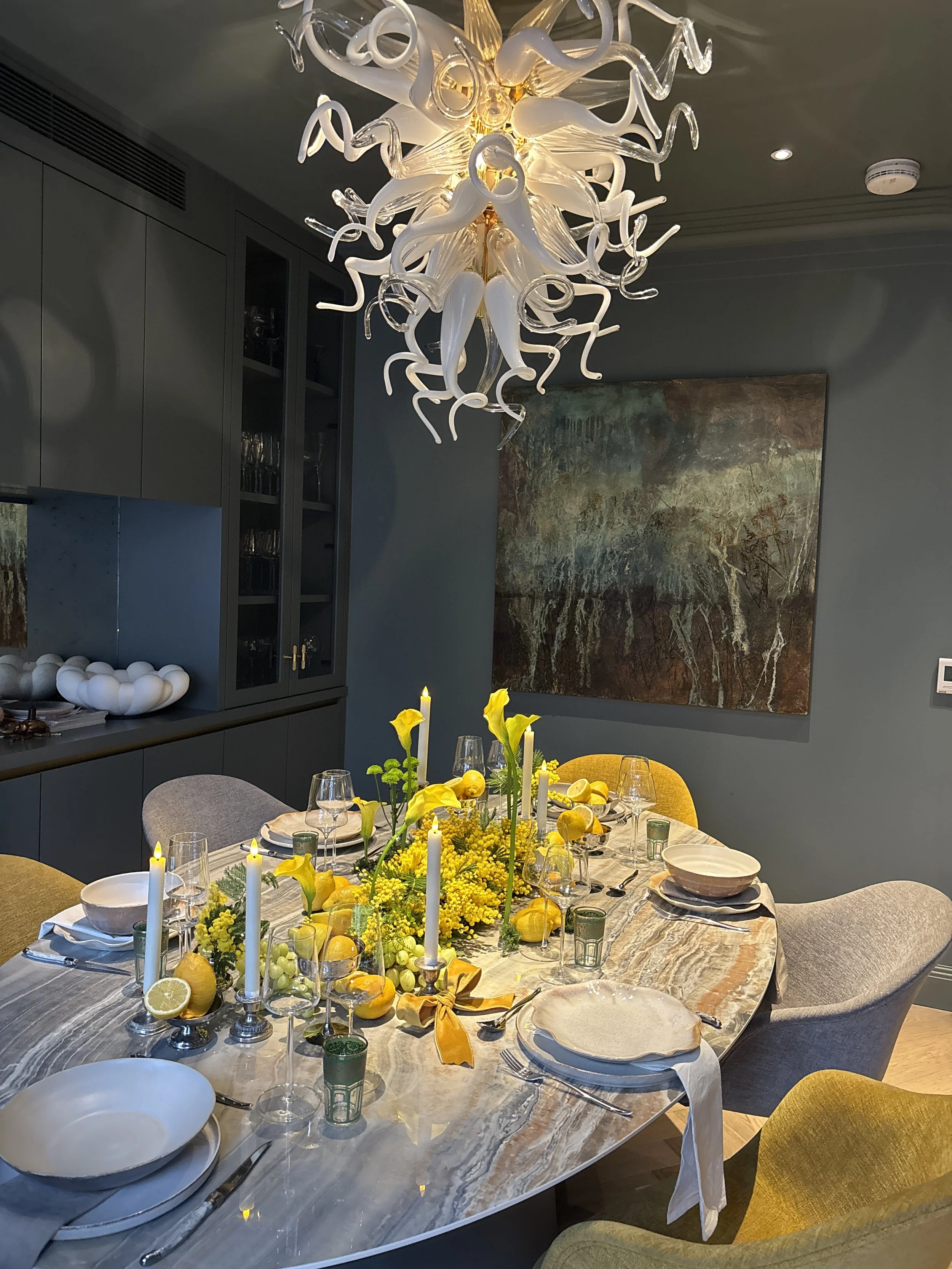 A dining room with a marble table set with plates, glasses, and cutlery, decorated with yellow flowers and candles. A chandelier hangs above, and the walls feature a large abstract painting. There are chairs around the table, some with yellow upholst