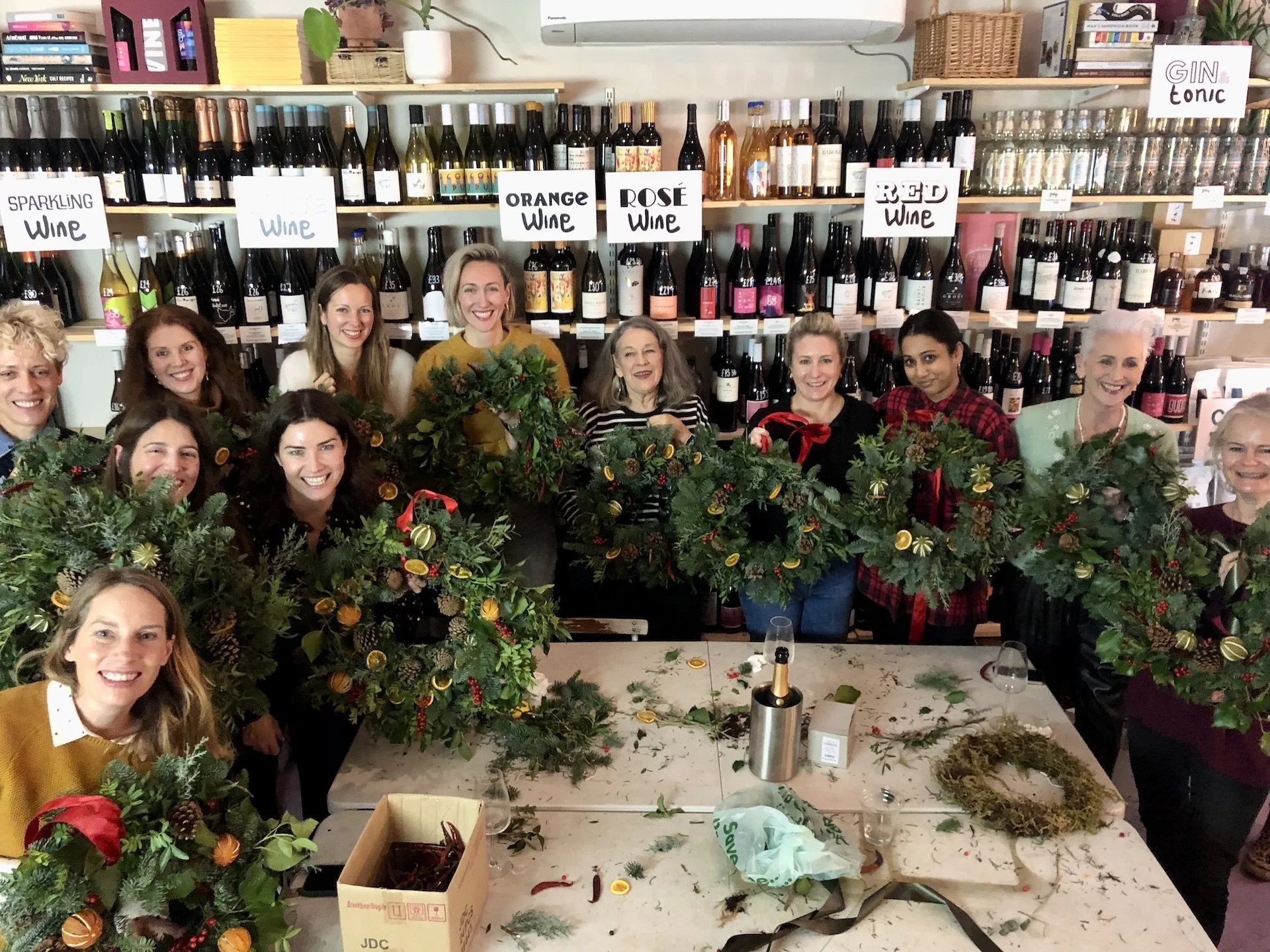 Group of women smiling and holding festive holiday wreaths decorated with oranges, pinecones, and ribbons, in a room with shelves of wine bottles and books in the background.