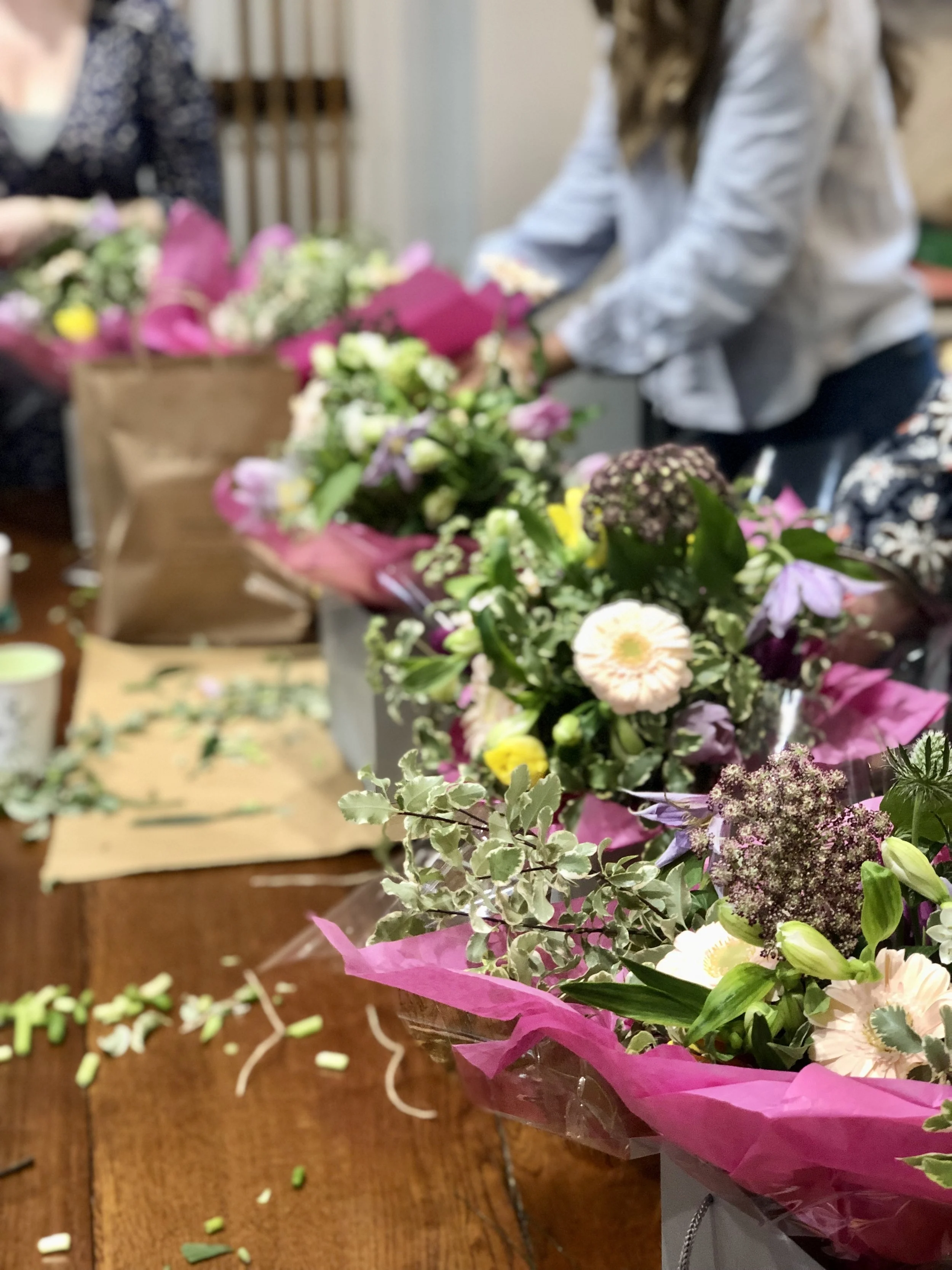 Close-up of colorful flower bouquets wrapped in pink paper on a wooden table, with some loose flower parts and greenery around.