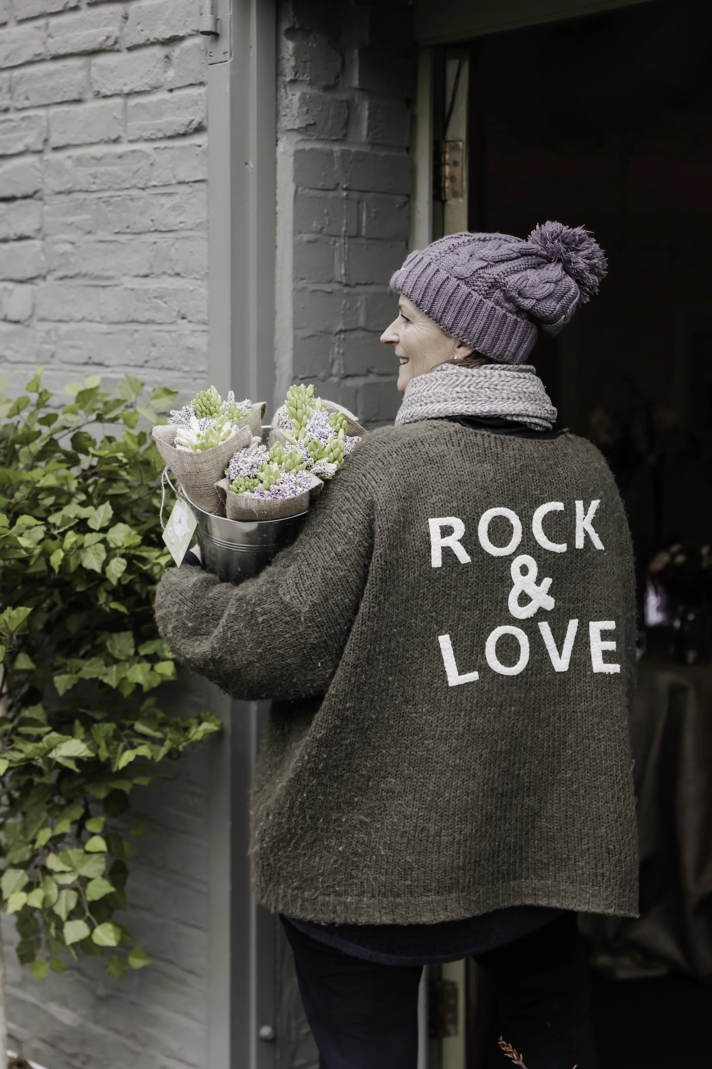 A smiling woman wearing a purple knit hat with a pom-pom, a gray scarf, and a brown sweater with the word 'ROCK & LOVE' on the back, holding a bouquet of flowers near a brick building.