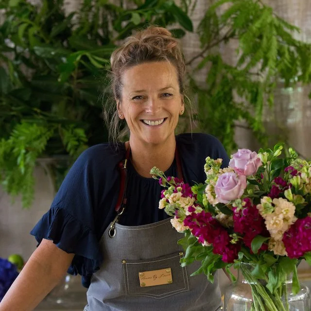 A woman with curly hair tied back, smiling, wearing a dark blue top and apron, standing behind a bouquet of pink and cream flowers, with green plants in the background.