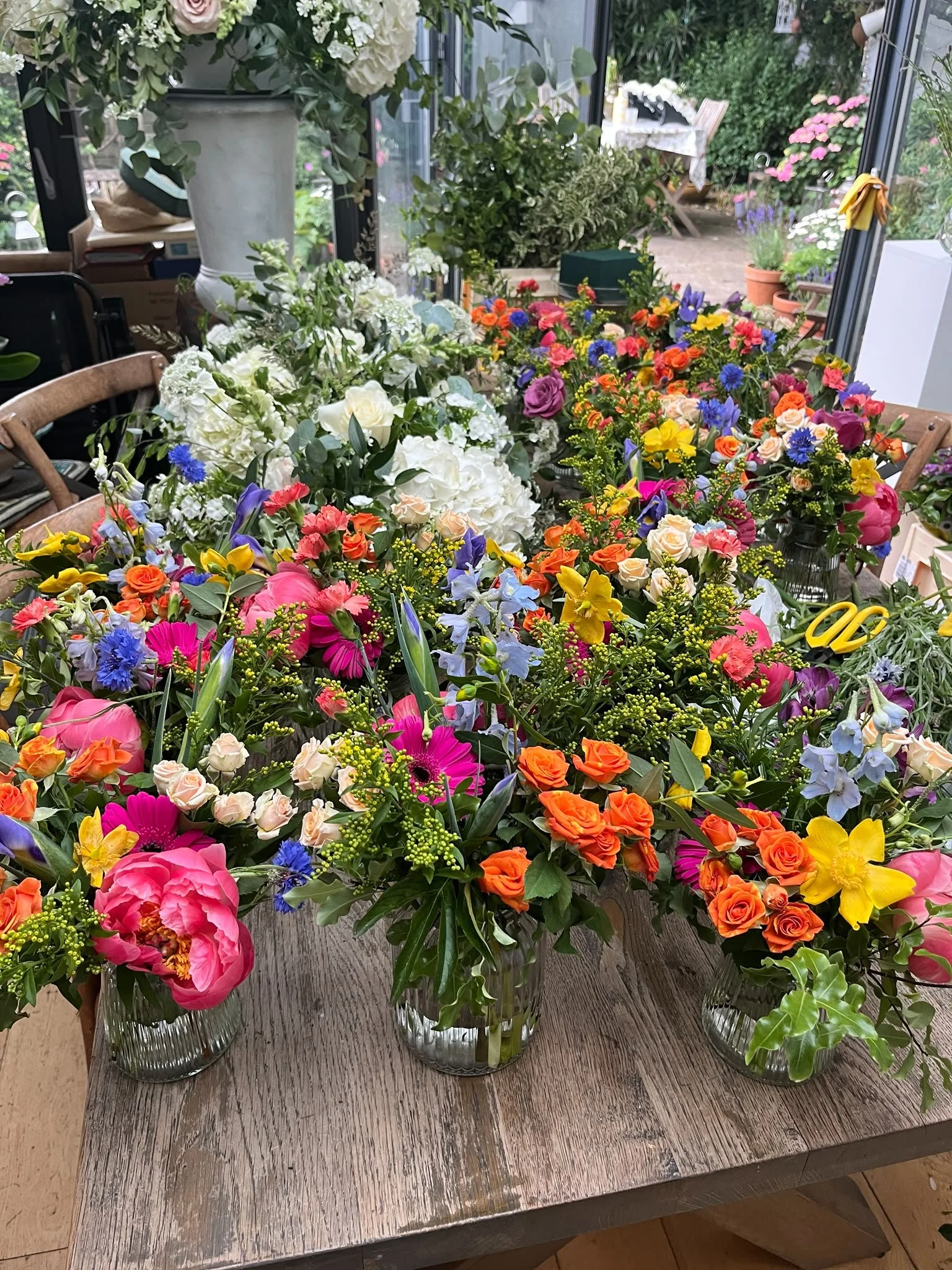 Various colorful flower arrangements in glass vases on a rustic wooden table, with a garden visible in the background.
