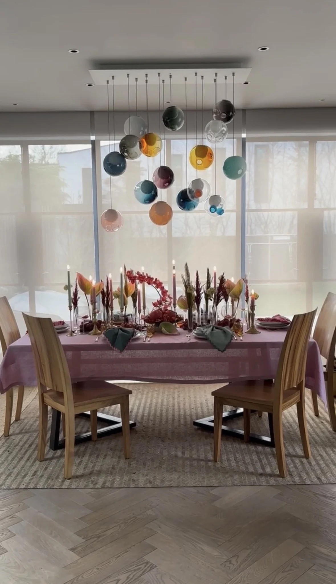 A dining table set for a celebration or dinner, decorated with pink tablecloth, floral centerpieces, candles, and placemats. Hanging from the ceiling above the table are colorful glass orbs for decoration. Large windows with sheer curtains provide na